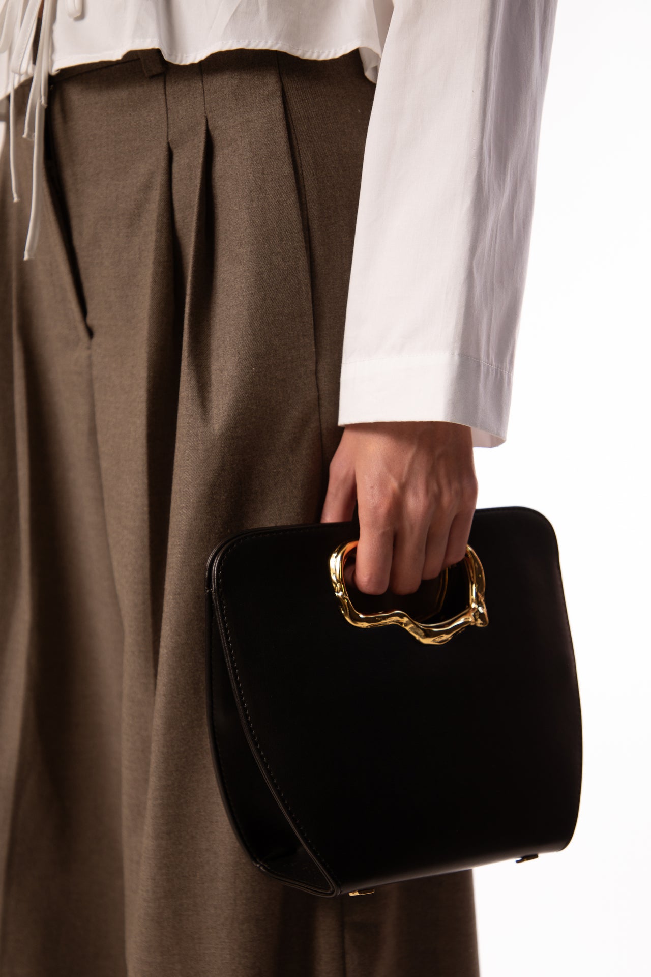 A minimalist, black leather handbag with a sculptural gold-tone handle opening, featuring an asymmetrical organic design.