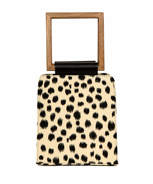 A modern animal-print handbag with a structured silhouette, black brushstroke-style spots, and a square wooden handle with a black leather accent.