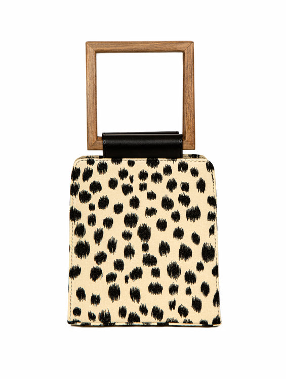 A modern animal-print handbag with a structured silhouette, black brushstroke-style spots, and a square wooden handle with a black leather accent.