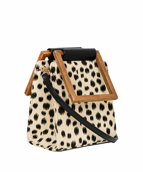 A stylish, boxy handbag with a cream-colored base and irregular black leopard spots, complemented by a geometric wooden handle