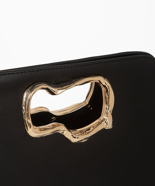 A minimalist, black leather handbag with a sculptural gold-tone handle opening, featuring an asymmetrical organic design.