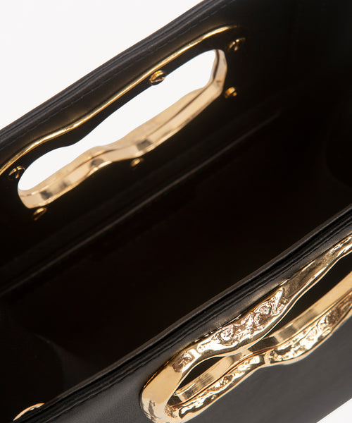 A minimalist, black leather handbag with a sculptural gold-tone handle opening, featuring an asymmetrical organic design.