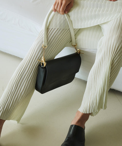 Elegant black shoulder bag with unique gold hardware and contrasting white handles, styled with a pleated cream outfit and black mules for a sophisticated, modern look