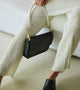 Elegant black shoulder bag with unique gold hardware and contrasting white handles, styled with a pleated cream outfit and black mules for a sophisticated, modern look
